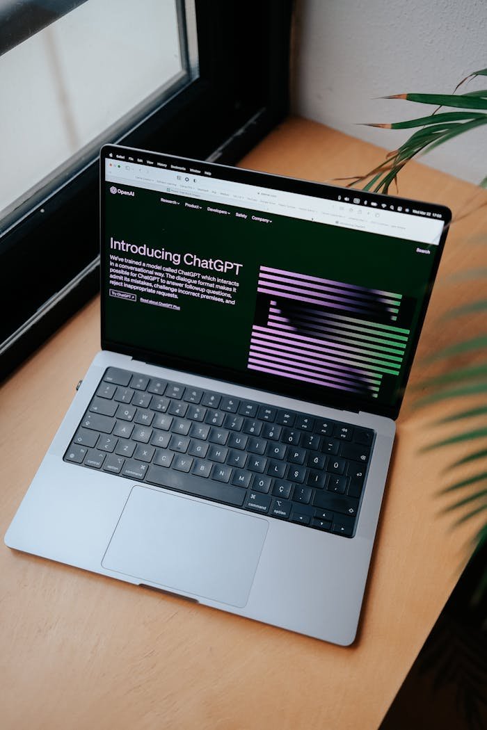 A sleek laptop on a wooden desk showing the ChatGPT homepage by a window.